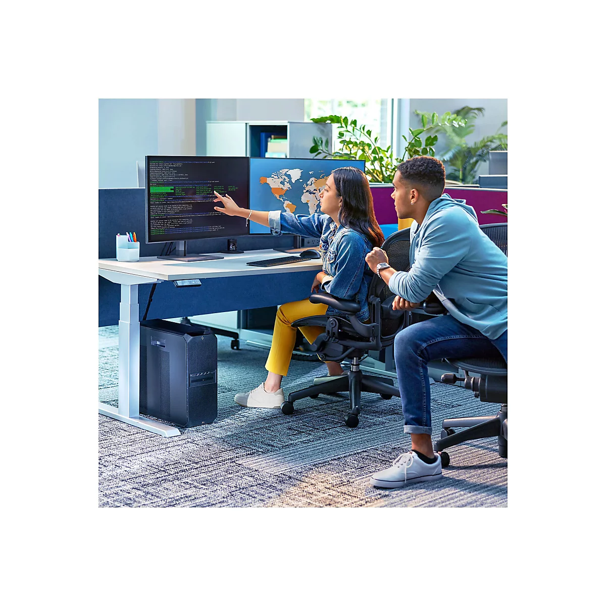 Zwei Personen sitzen an einem Schreibtisch, der an einem Computer arbeitet. Eine Person zeigt auf den Bildschirm, der Code anzeigt. Die andere Person schaut zu.