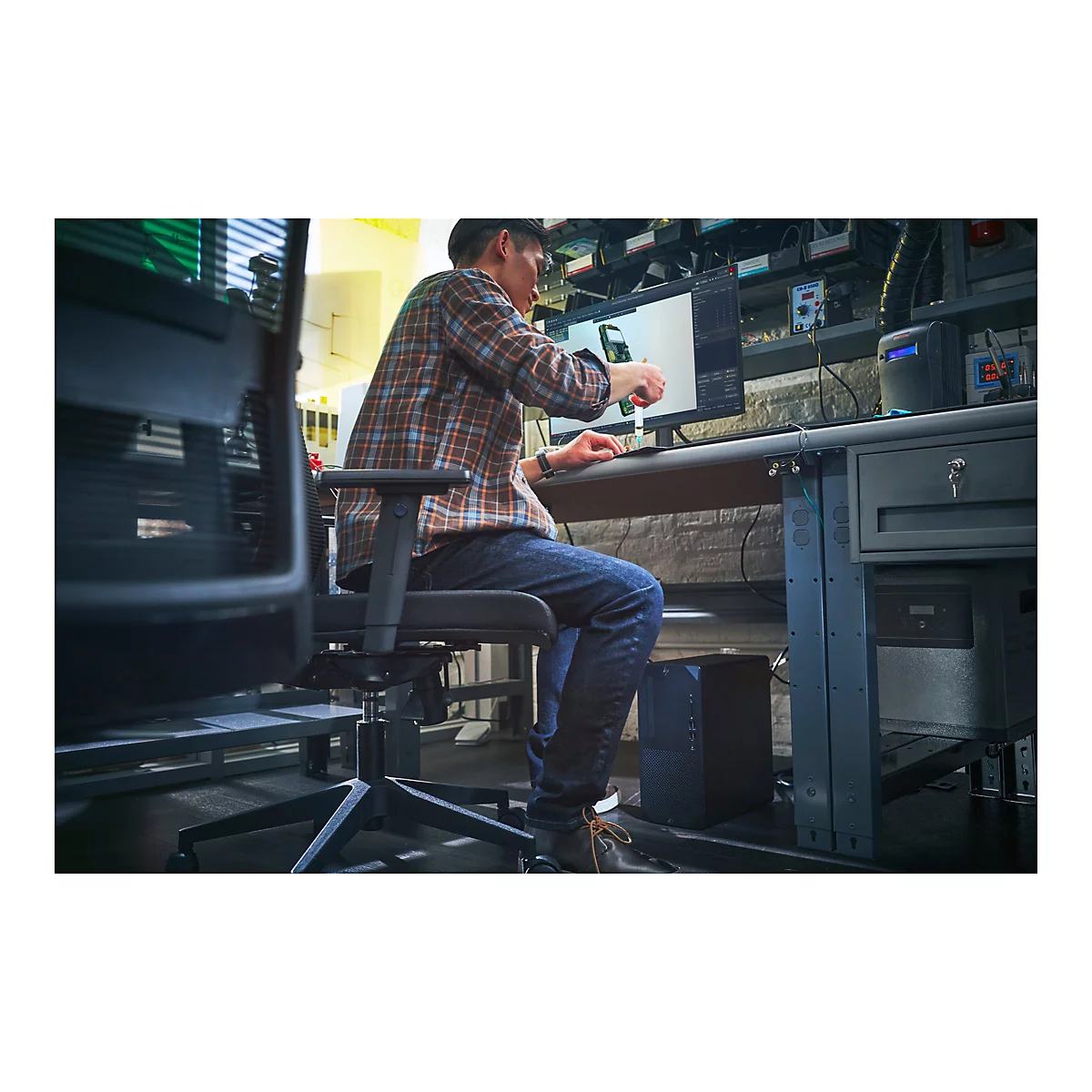 Eine Person sitzt an einem Schreibtisch und arbeitet an einem Computer. Er trÃ¤gt ein kariertes Hemd und Jeans. Der Hintergrund zeigt technische GerÃ¤te.
