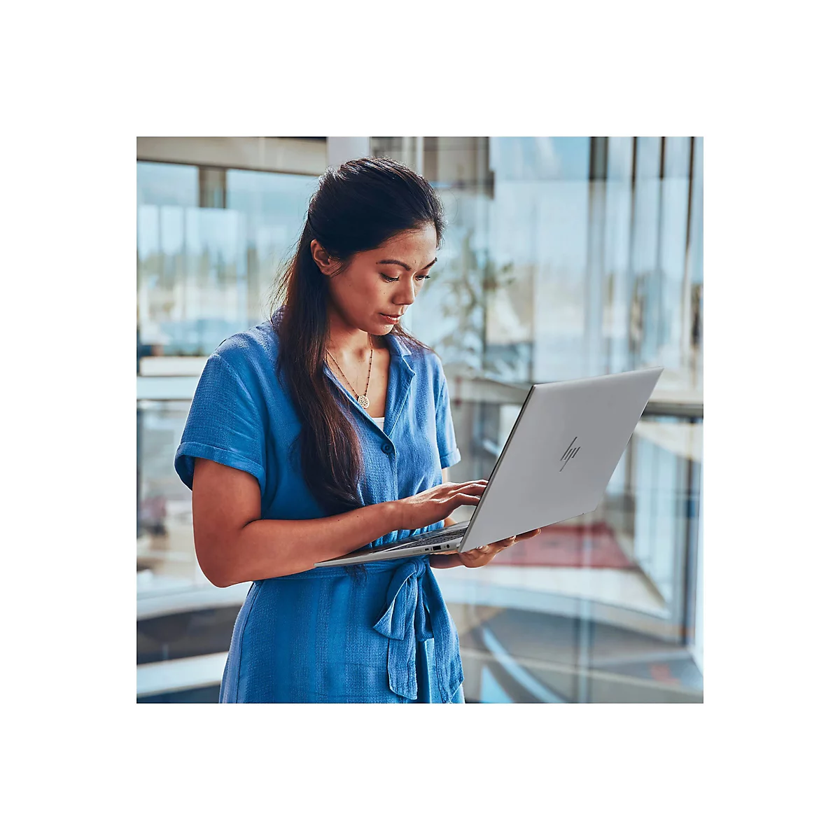 Eine Frau in blauem Kleid arbeitet an einem Laptop. Sie steht vor einem Fenster mit Blick auf eine urbane Umgebung.