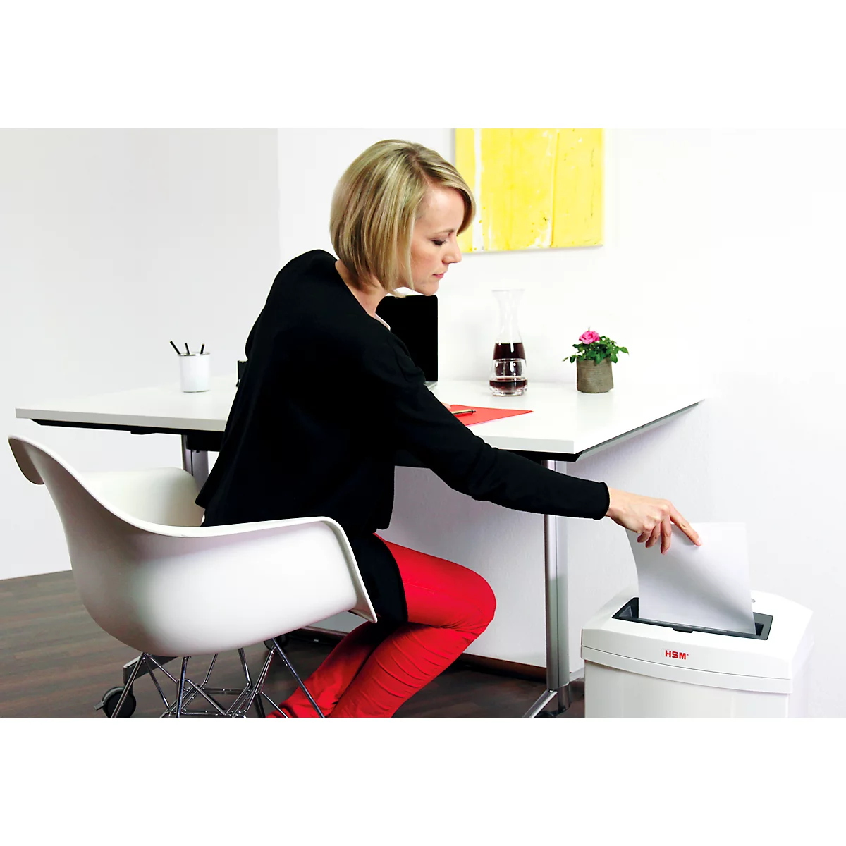 A woman in red tights is putting paper into a shredder. She sits at a desk with a laptop.