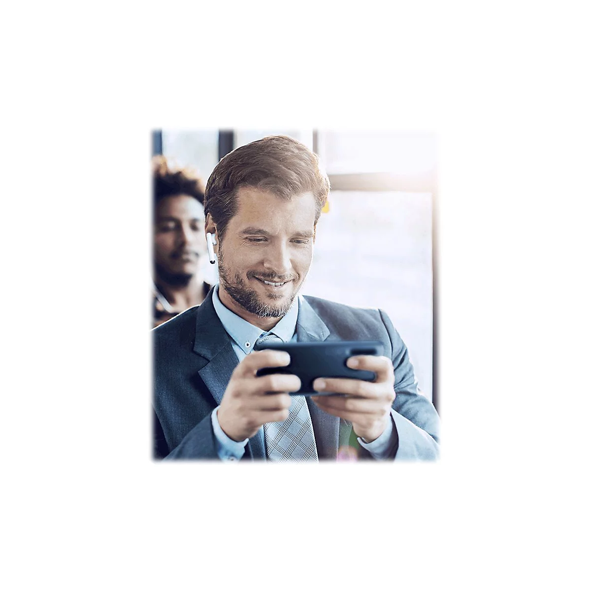Ein lächelnder Mann im Anzug, mit kabellosen Kopfhörern, blickt auf sein Smartphone. Eine Person ist im Hintergrund unscharf.