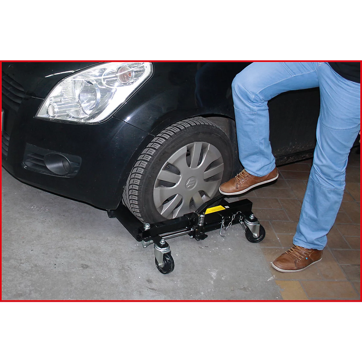 Ein Mann in Jeans und braunen Schuhen steht neben dem Vorderrad eines schwarzen Autos, das auf einem schwarzen Rangierwagen steht.