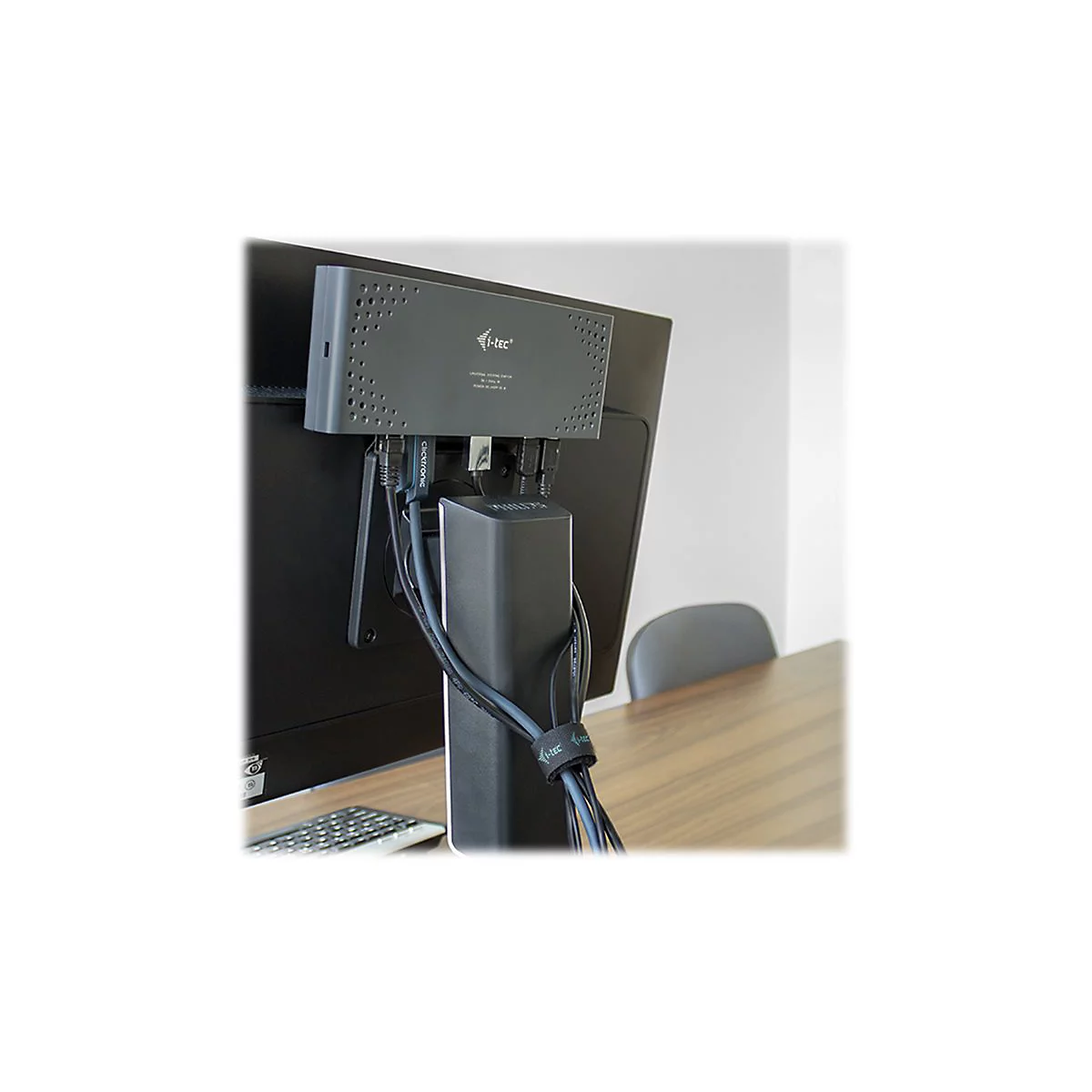 Adaptateur gris fixÃ© Ã  un moniteur, cÃ¢bles regroupÃ©s. Clavier et chaise visibles sur la table.