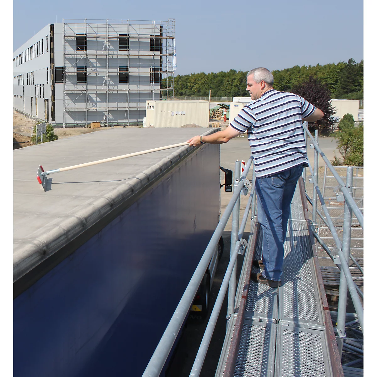 A man cleaning the roof of a blue trailer with a broom and a work platform.