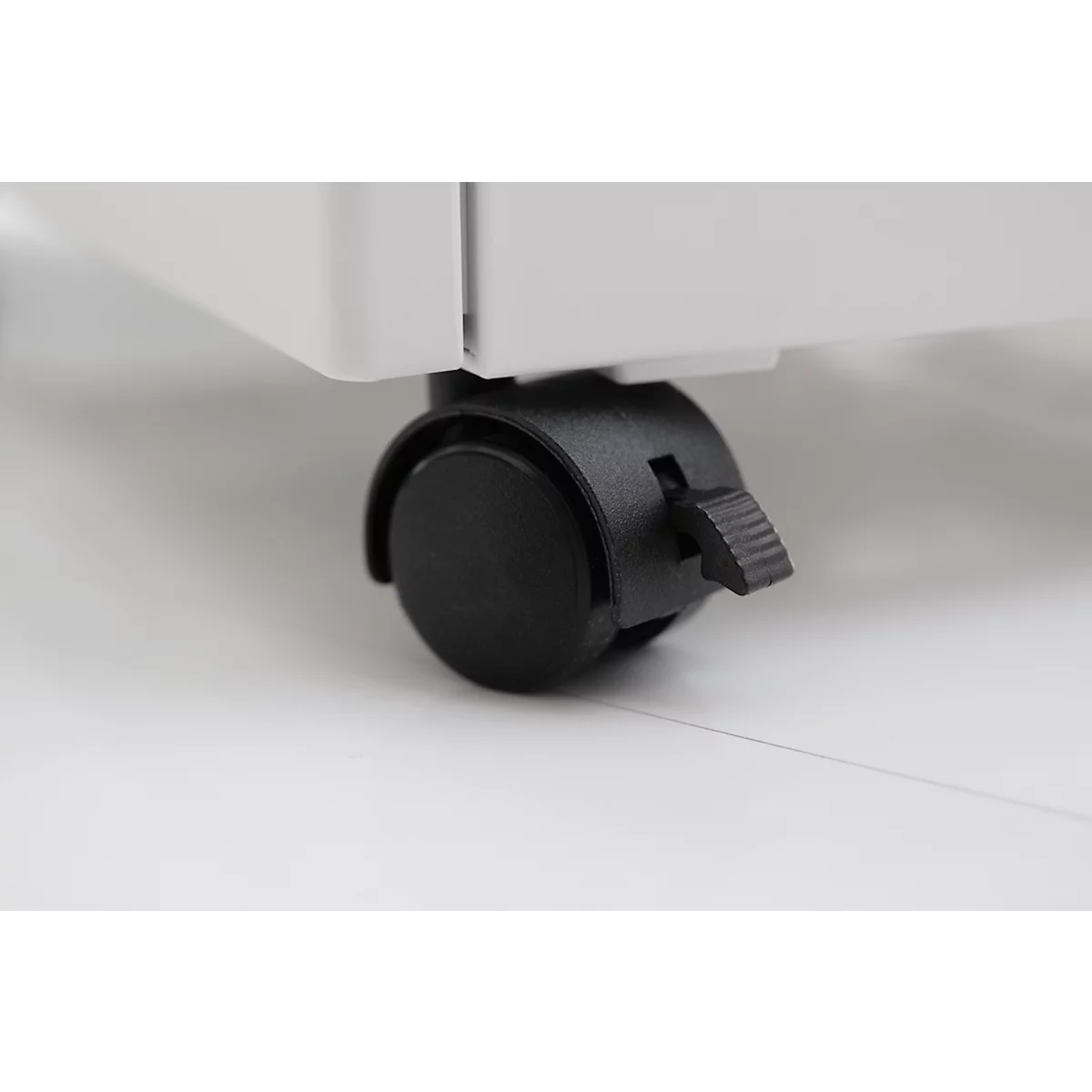 Nahaufnahme eines schwarzen Rollenrades unter einem weißen Möbelstück, mit schwarzem Feststellhebel. Der Untergrund ist weiß.