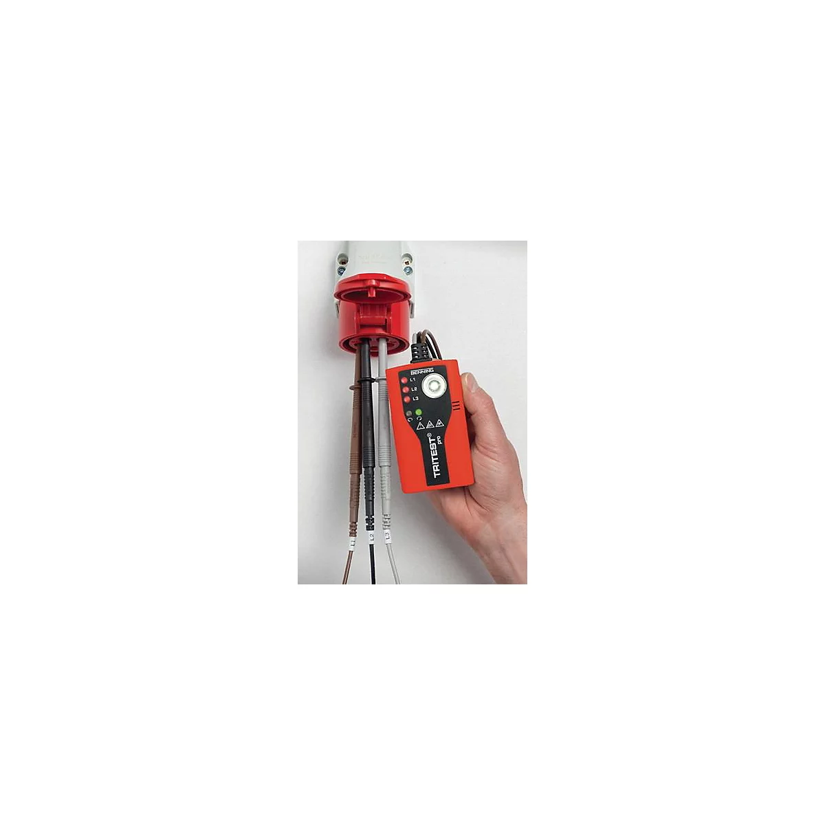 Un appareil de test TRITEST Pro rouge est tenu à la main tout en étant connecté à une prise rouge. Des câbles sont attachés.