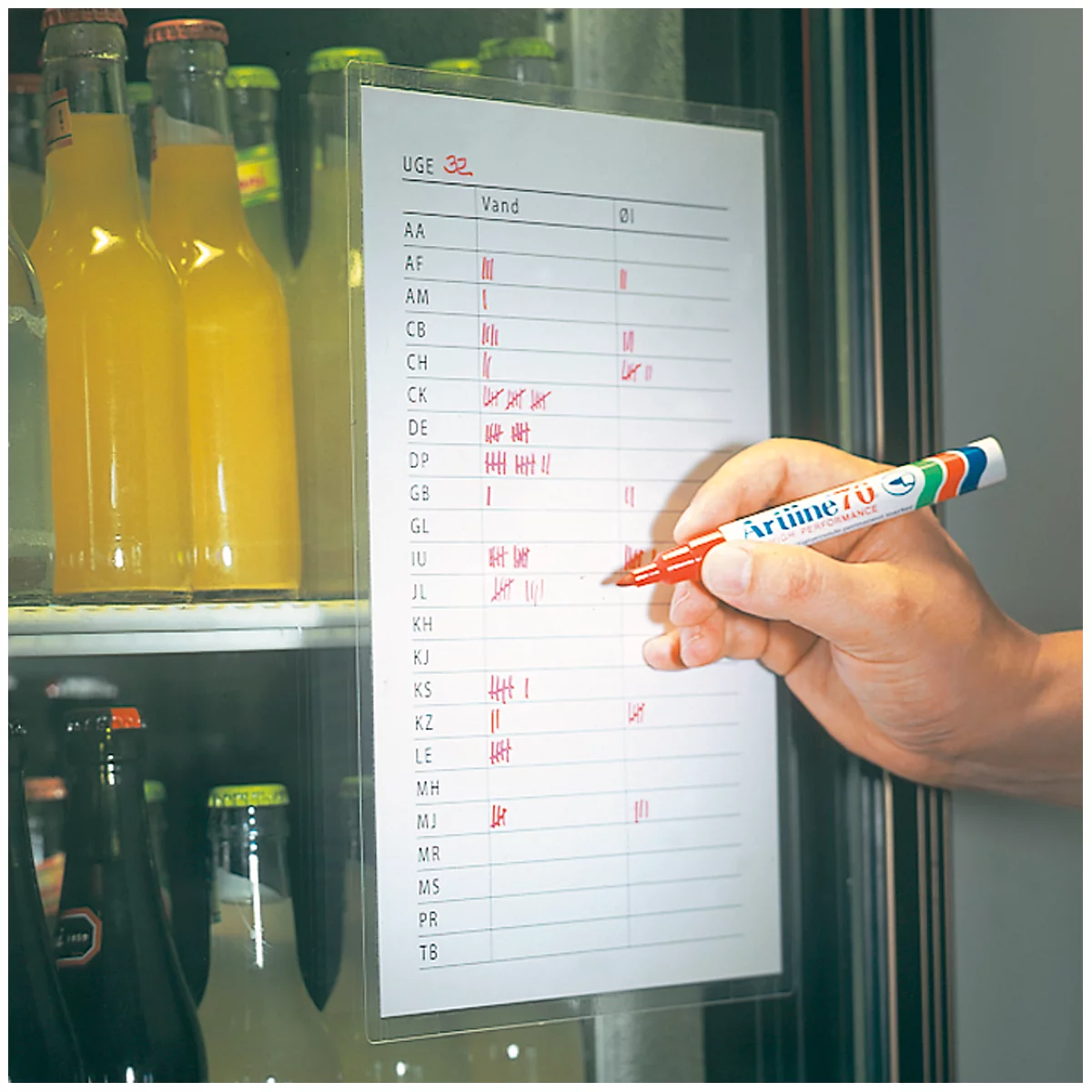 A man with a red pen writes on a list attached to a refrigerator door. Drinks in bottles are visible on the left.