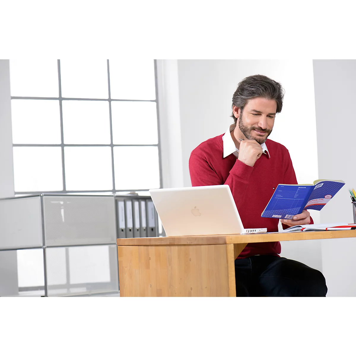 A man in a red sweater is reading at a desk in an office. There is a laptop and a book on the table. Large windows in the background.