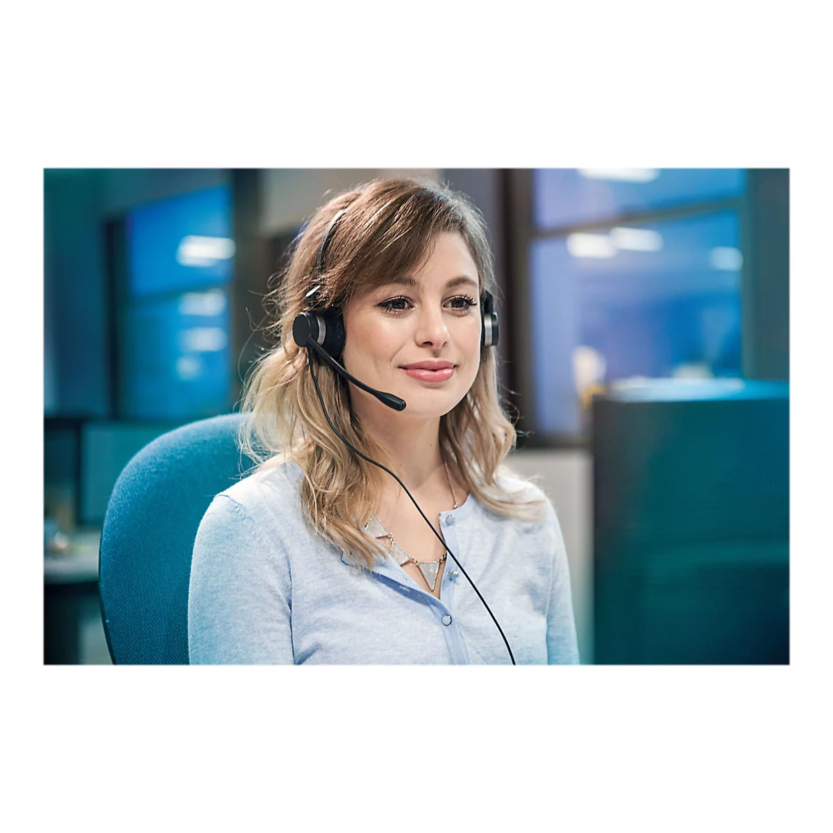 Eine Frau mit Headset, Blick auf den Monitor. Sie lÃ¤chelt leicht. Blaue Kleidung und Stuhl.