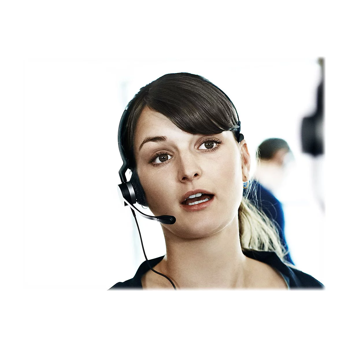 Junge Frau mit Headset, sie spricht oder hÃ¶rt zu. Im Hintergrund weitere Person unscharf.