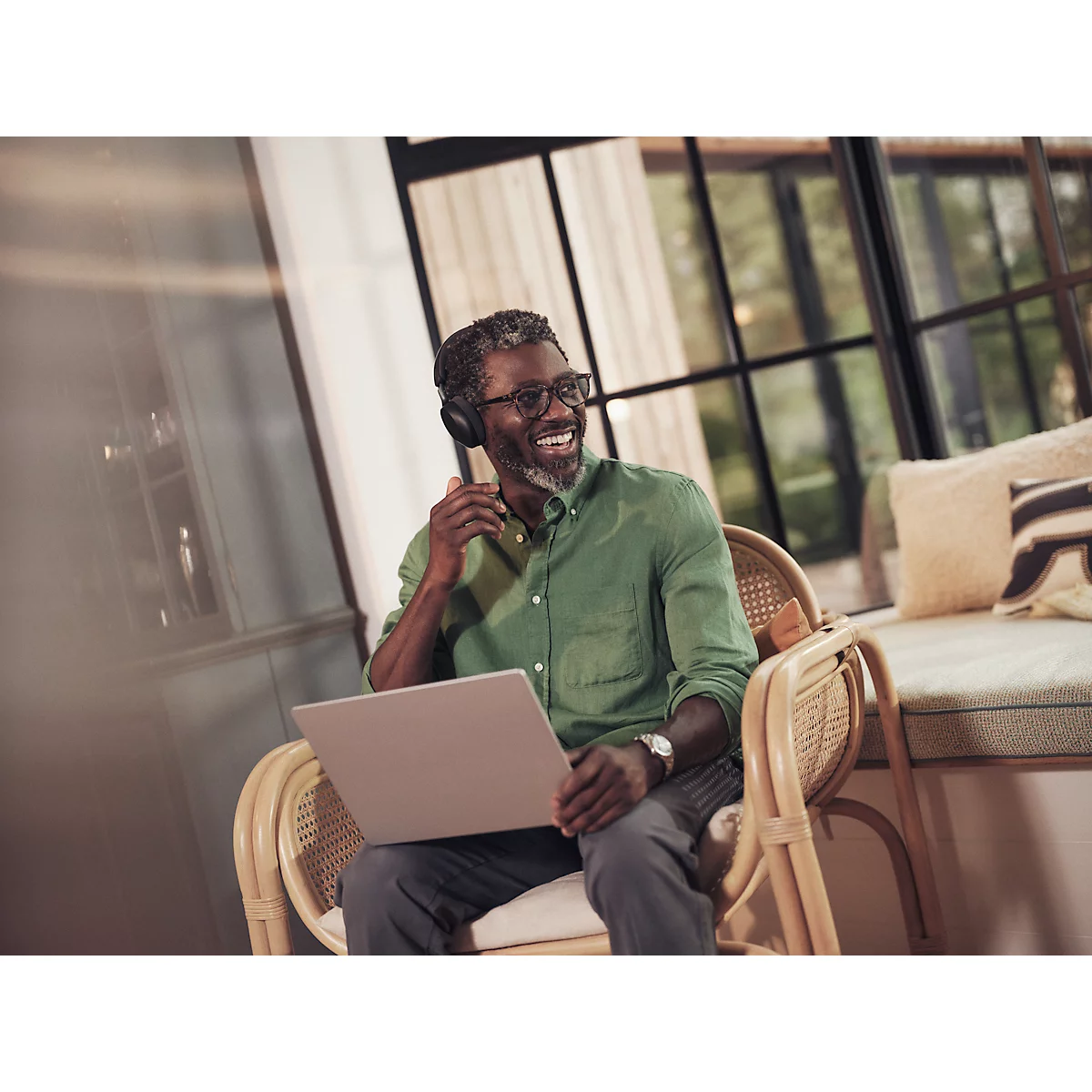 Un homme souriant est assis dans un fauteuil en rotin avec un ordinateur portable et un casque, des fenêtres et des oreillers en arrière-plan.