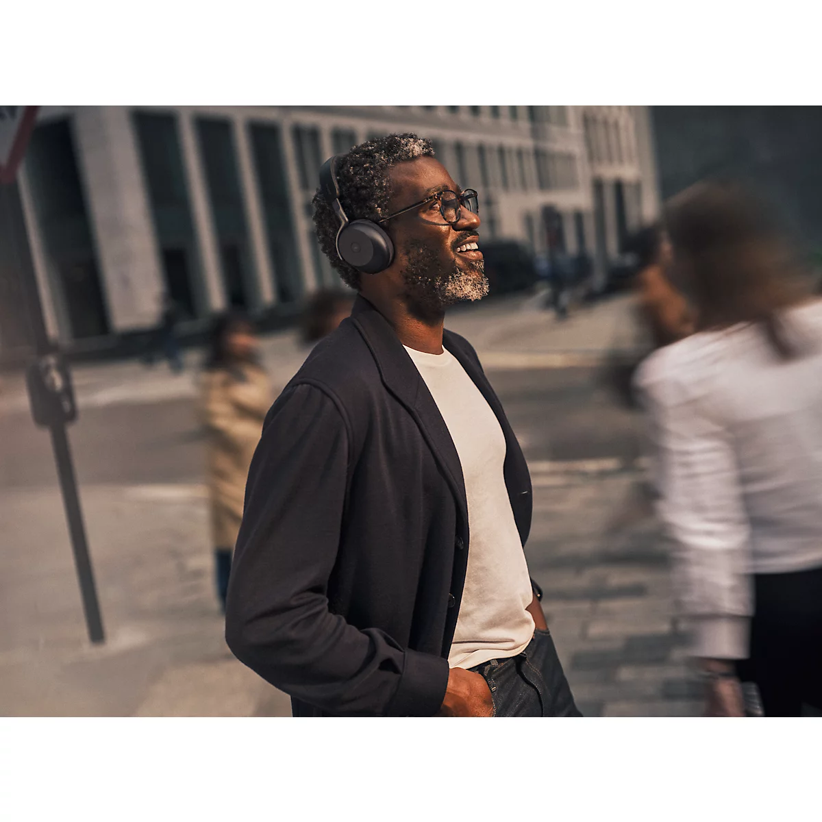 Un homme souriant avec un casque traverse une rue. Il porte des lunettes, une veste et une chemise blanche.