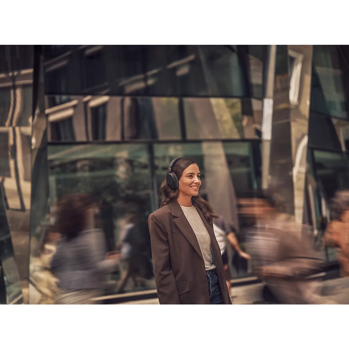 Une femme souriante portant un casque marche dans une ville. Elle porte un blazer marron et regarde vers la droite.