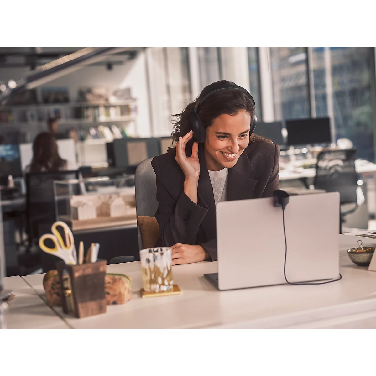 Une femme souriante avec un casque parle sur un ordinateur portable dans un bureau. Ciseaux, stylos et une boisson sont sur la table.