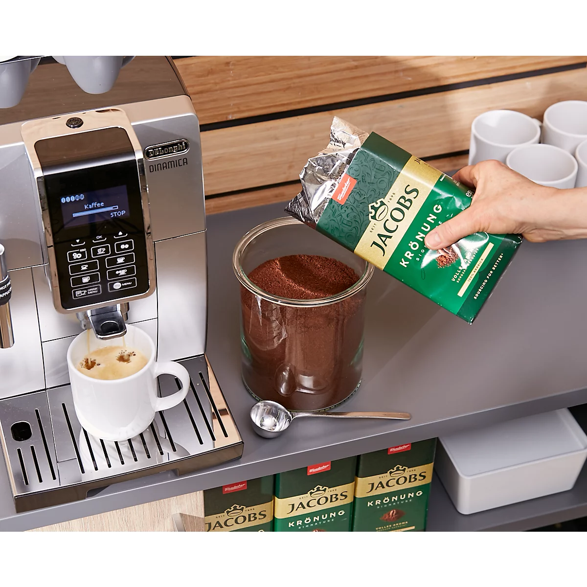 Coffee machine brewing coffee, next to it a hand pouring coffee powder into a glass jar. The bag reads "Jacobs Krönung".