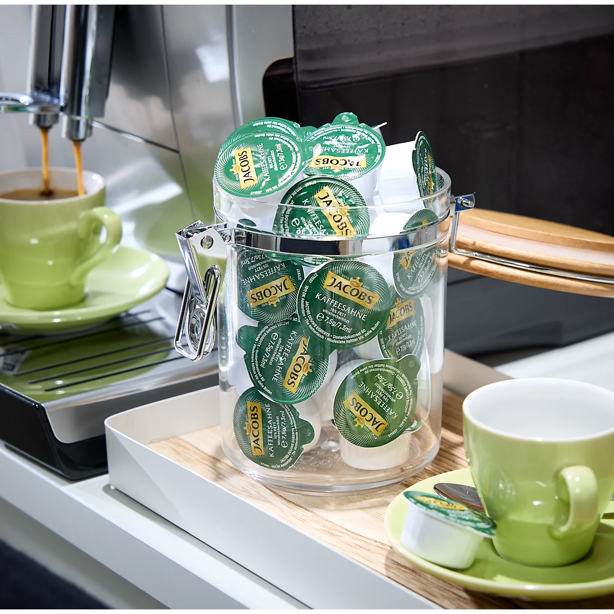 A coffee machine brewing coffee into a green cup, next to a jar of Jacobs coffee capsules.