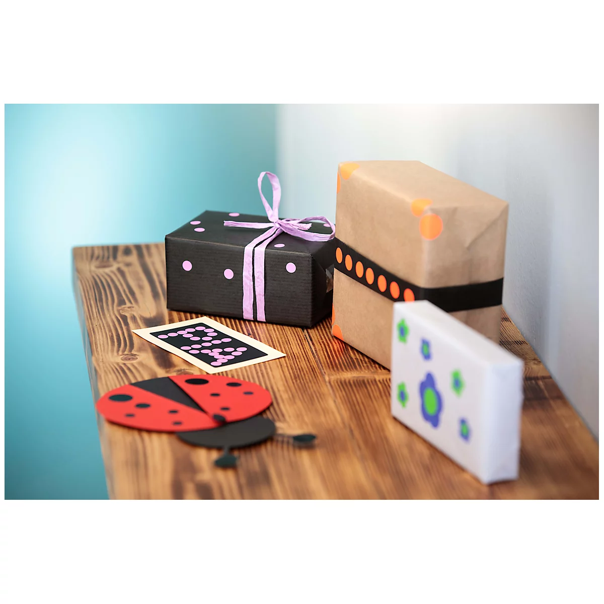 Trois boîtes cadeaux sont posées sur une table en bois. Une boîte noire avec des pois violets et un ruban. Une boîte marron avec une bande noire. Une boîte blanche avec des fleurs bleues.