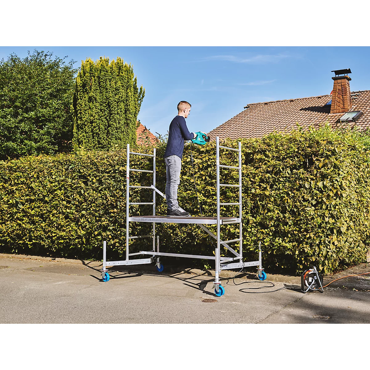 Un homme sur un échafaudage taille une haie. Une maison en arrière-plan.