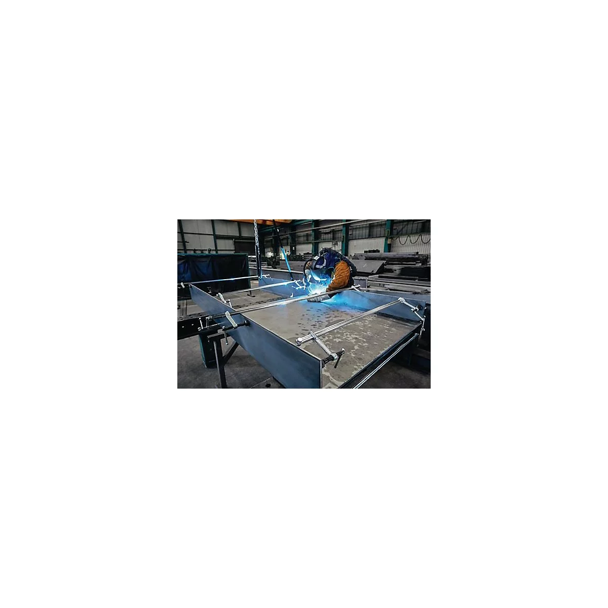 Un ouvrier soude des pièces métalliques dans une usine. Des étincelles bleues et des lunettes de protection sont visibles.