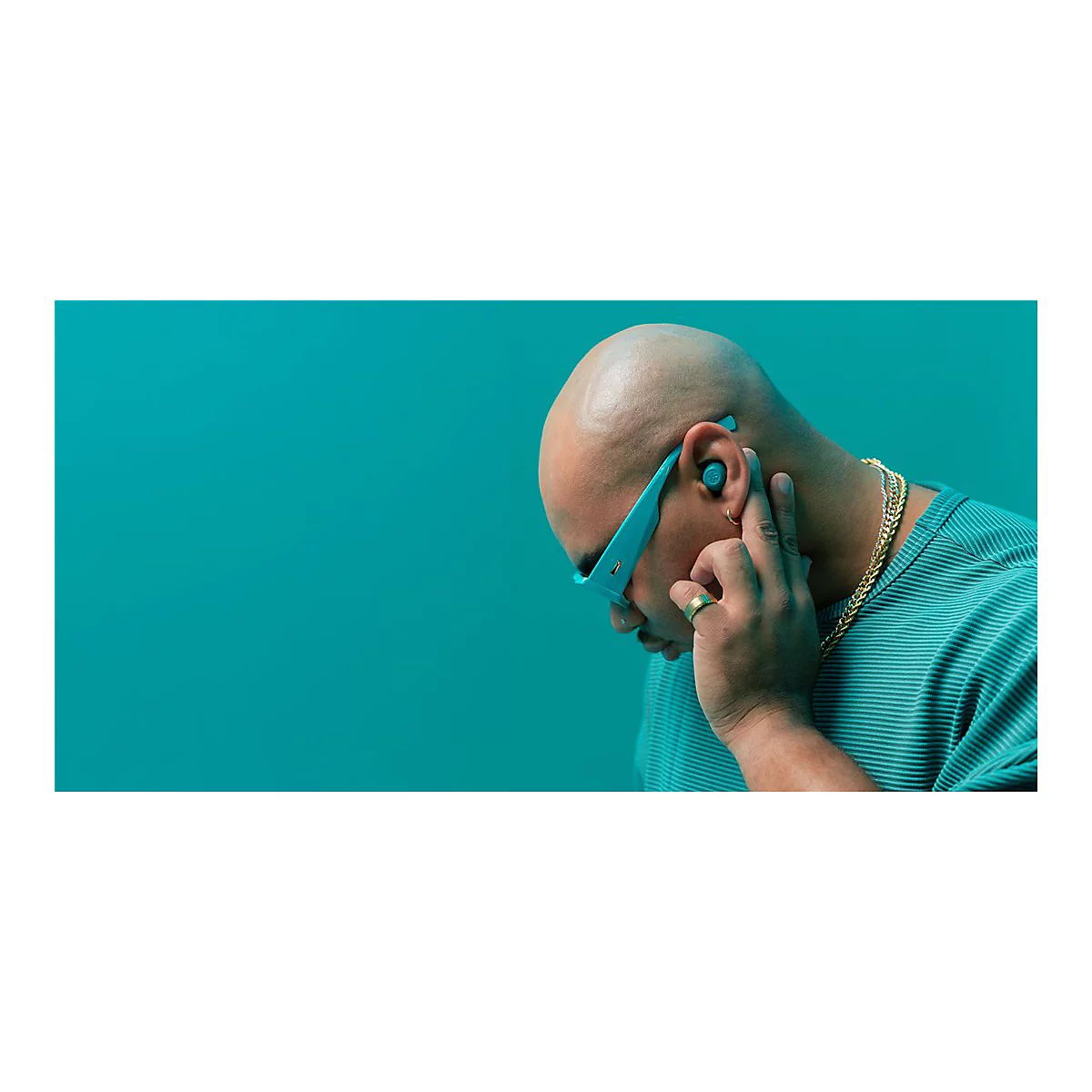 Un homme chauve portant un casque et des lunettes de soleil bleus touche son oreille, fond turquoise.