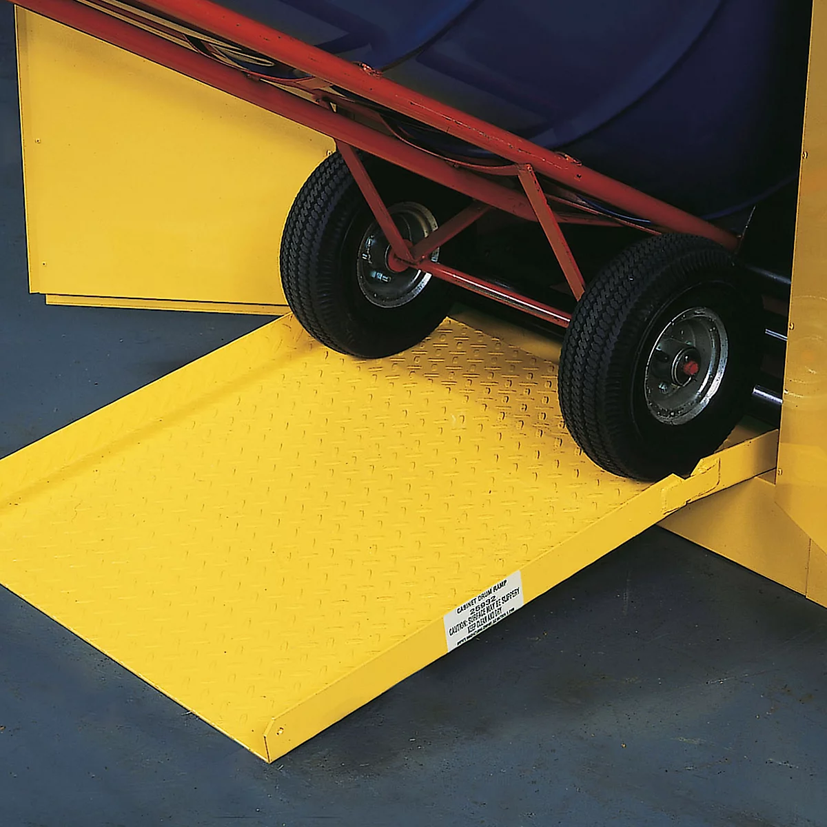 Yellow metal ramp for a trolley; two black tires stand on it. Text on the ramp.