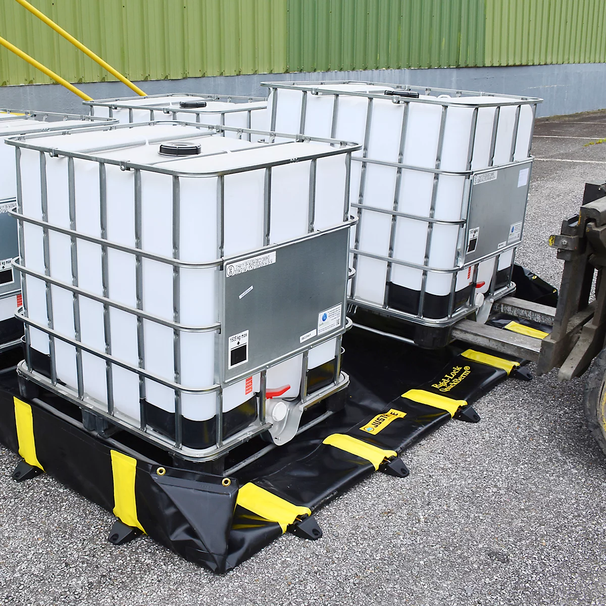 Verschillende witte IBC-containers op zwarte opvangbakken met gele accenten, op een geasfalteerd oppervlak.