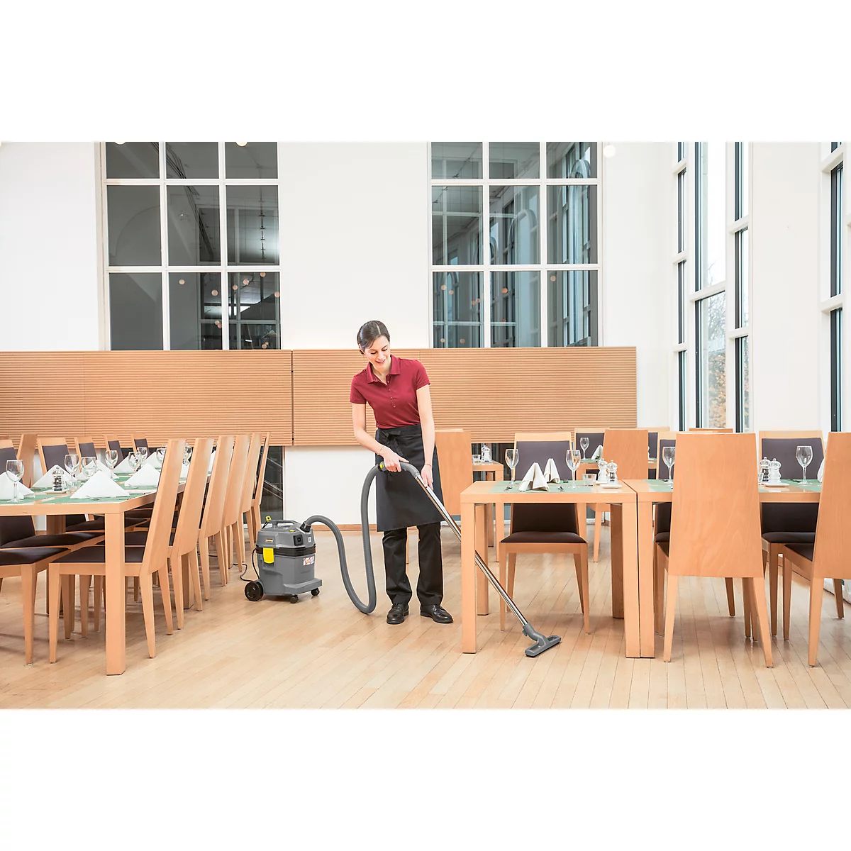 A waitress vacuums the floor in a restaurant, surrounded by tables, chairs, and windows.