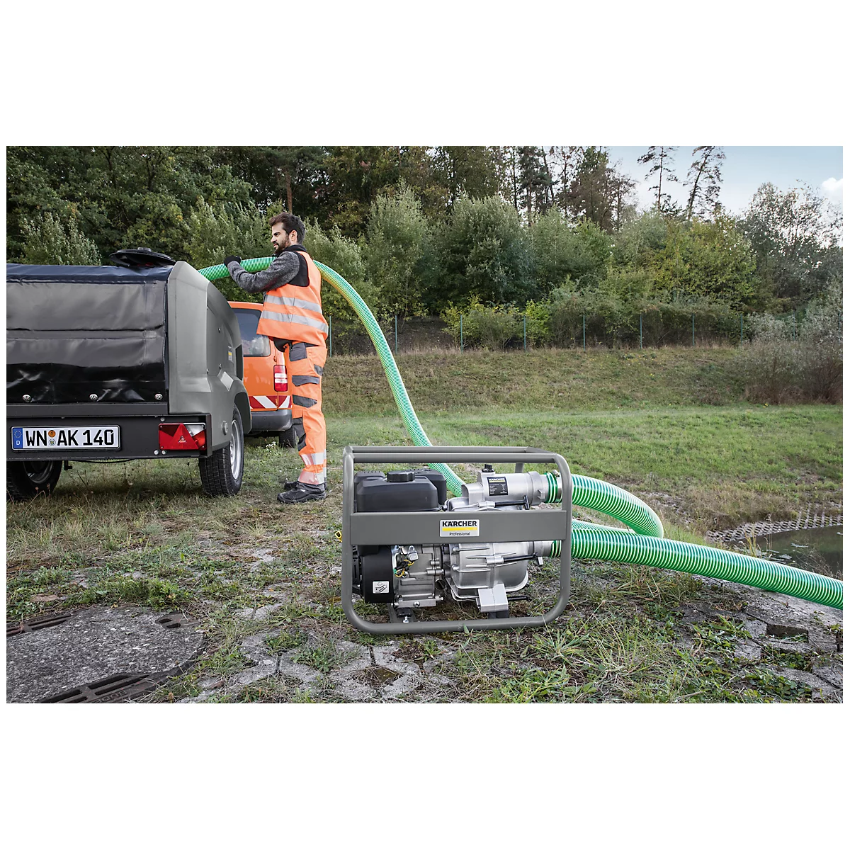 Man met pomp, trekt groene slang uit aanhanger, draagt veiligheidsvest, bomen op de achtergrond.