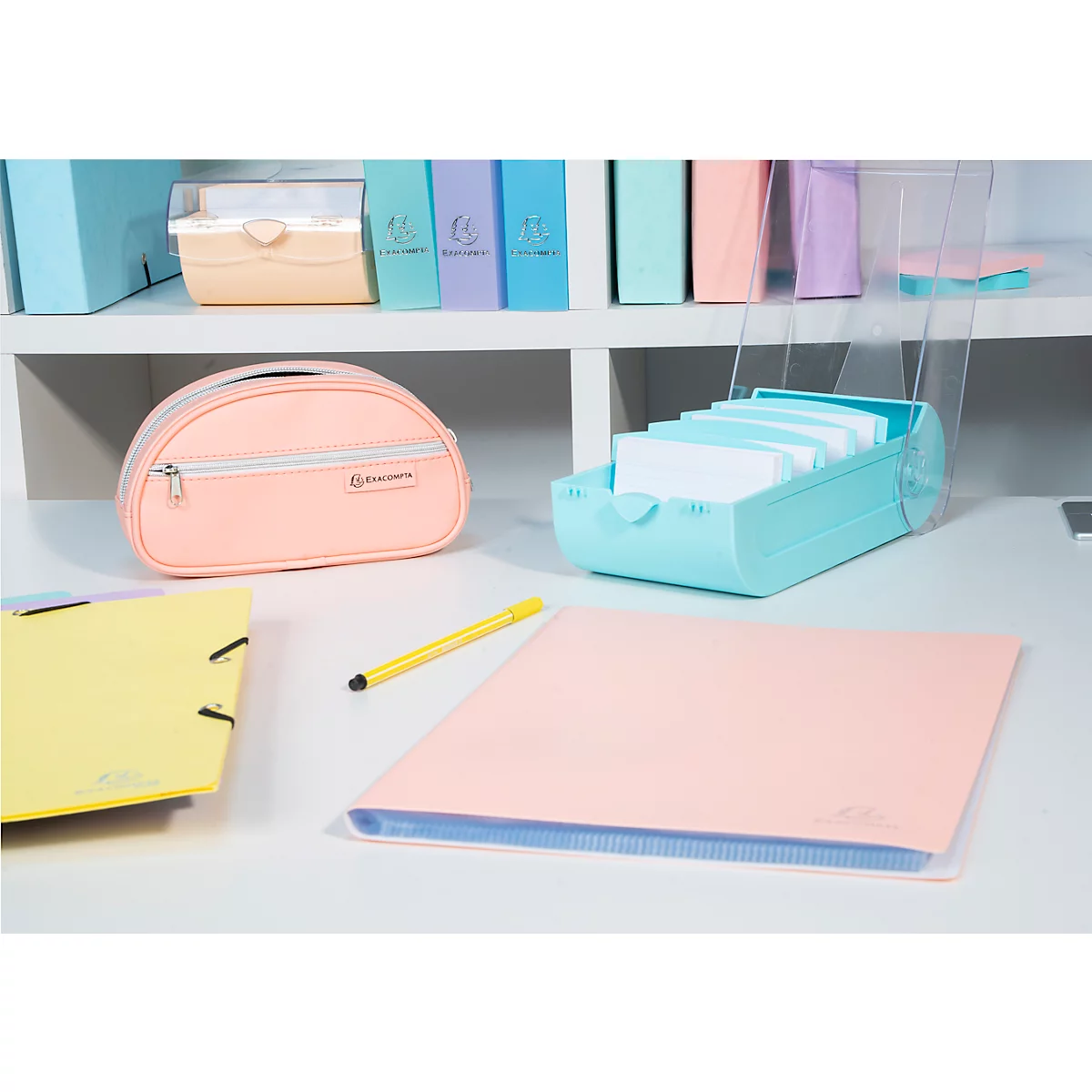 Divers articles de bureau sur un bureau : une trousse rose, un porte-stylos, un classeur rose, un document jaune et un stylo.