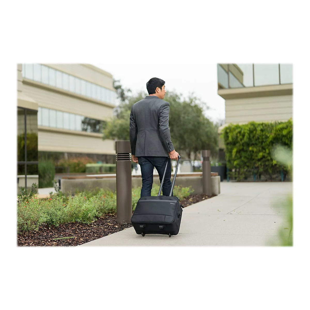 Ein Mann in einem Anzug zieht einen schwarzen Rollkoffer auf einem BÃ¼rgersteig. Im Hintergrund sind GebÃ¤ude und GrÃ¼nflÃ¤chen zu sehen.