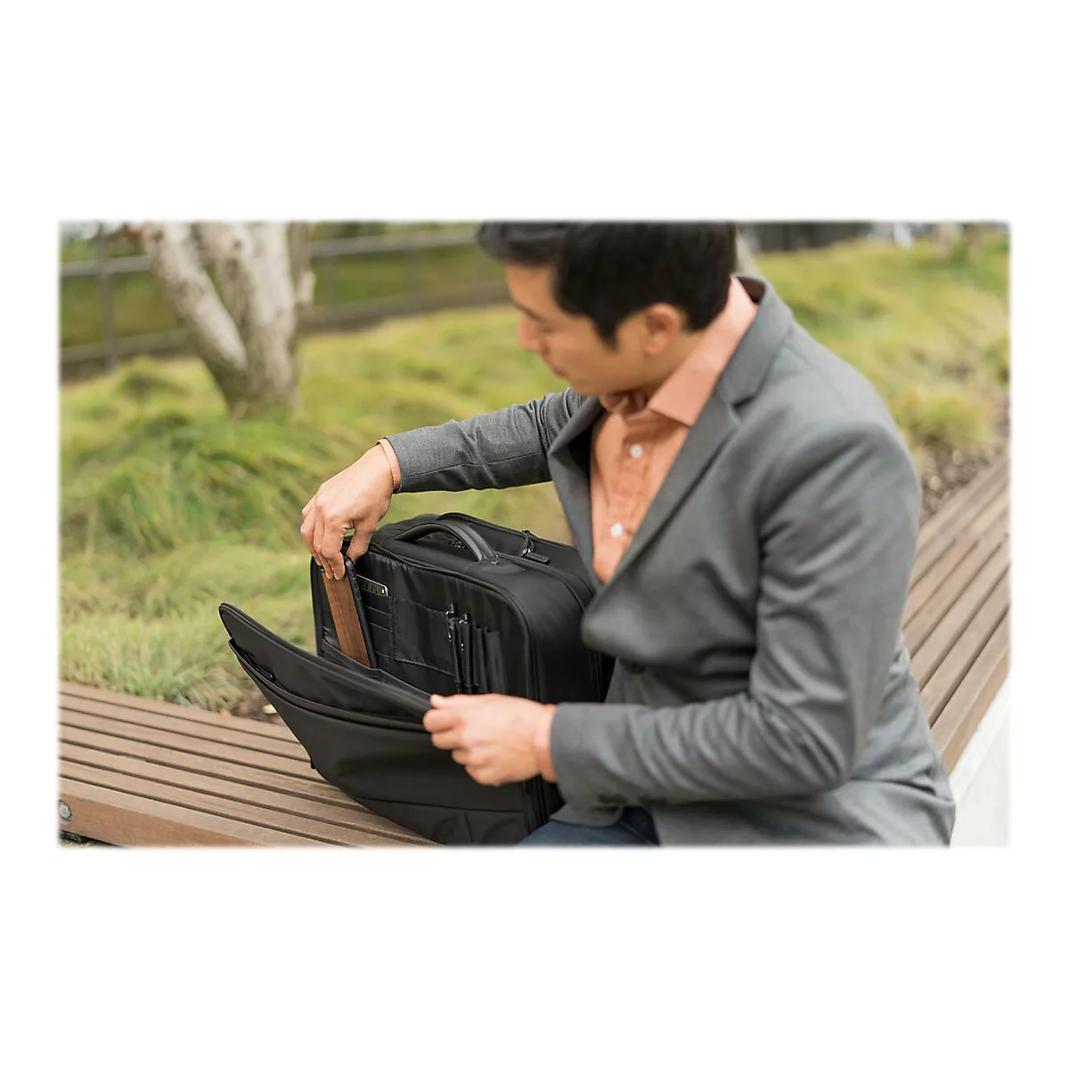 Ein Mann im Anzug Ã¶ffnet eine schwarze Aktentasche. Er sitzt auf einer Bank im Freien.