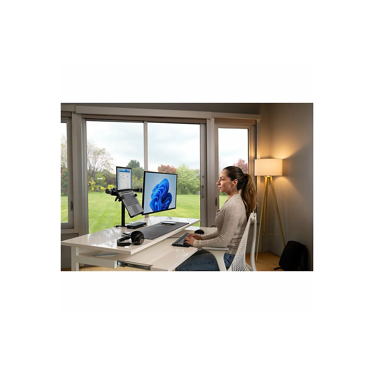 Femme à son bureau. Deux écrans, ordinateur portable, casque. Vue à travers la fenêtre sur le jardin.