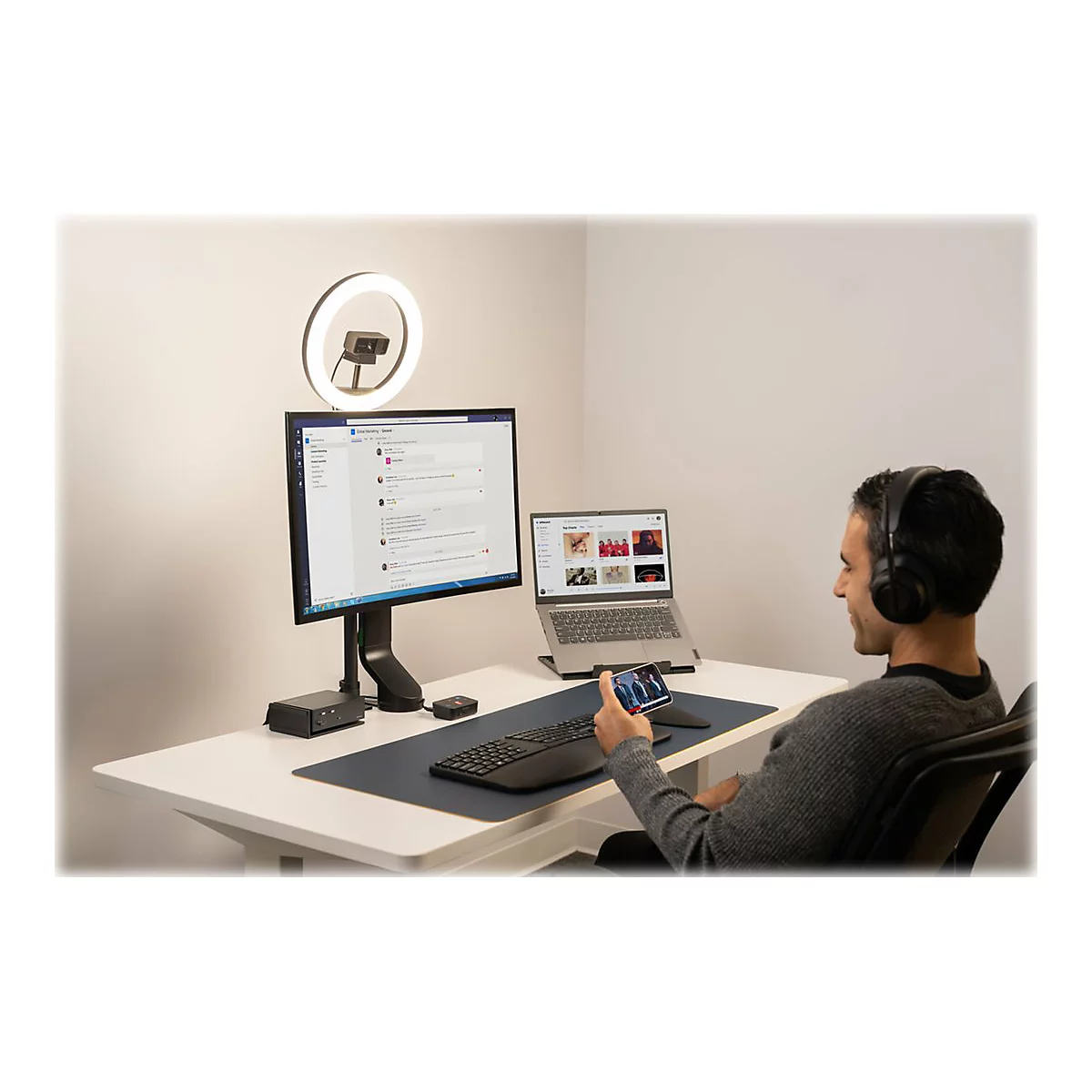 Homme avec un casque assis Ã  un bureau, utilisant un ordinateur portable, un moniteur et un smartphone. LumiÃ¨re annulaire et webcam Ã©galement prÃ©sentes.