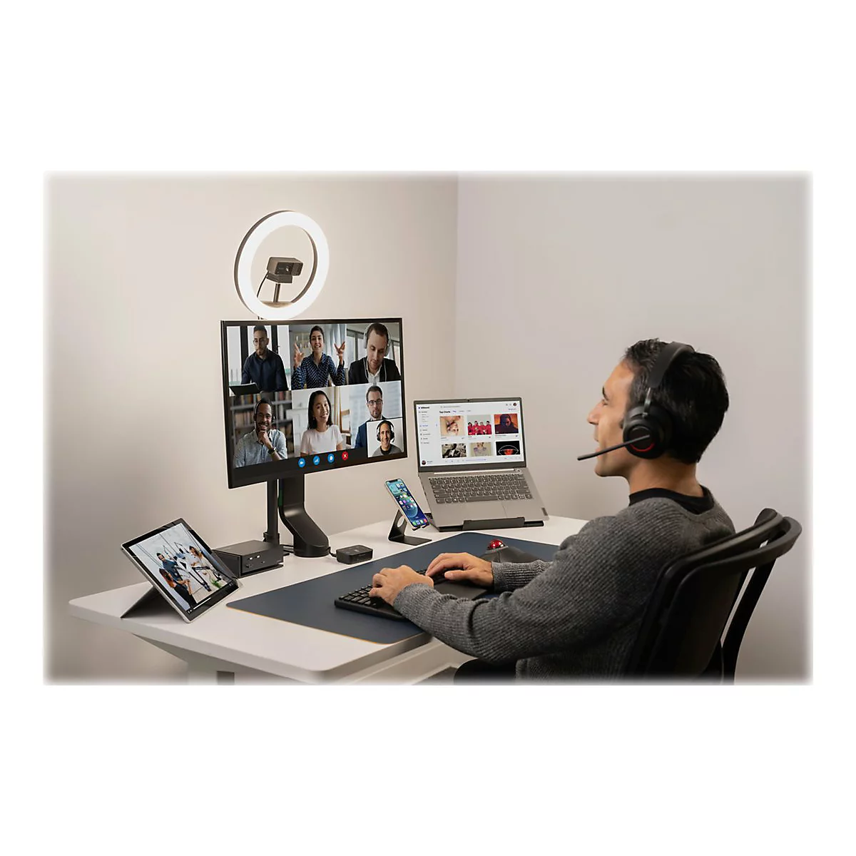 Un homme avec un casque participe Ã  une vidÃ©oconfÃ©rence. Sur la table : ordinateur portable, tablette, smartphone, anneau lumineux, moniteur.