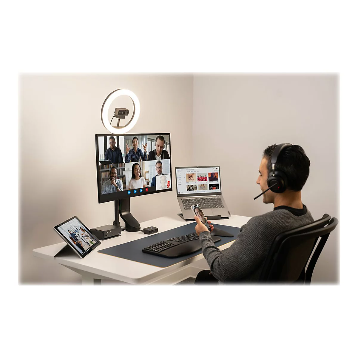 Un homme avec un casque participe Ã  une vidÃ©oconfÃ©rence. Sur le bureau : Ã©cran, tablette, ordinateur portable, anneau lumineux.