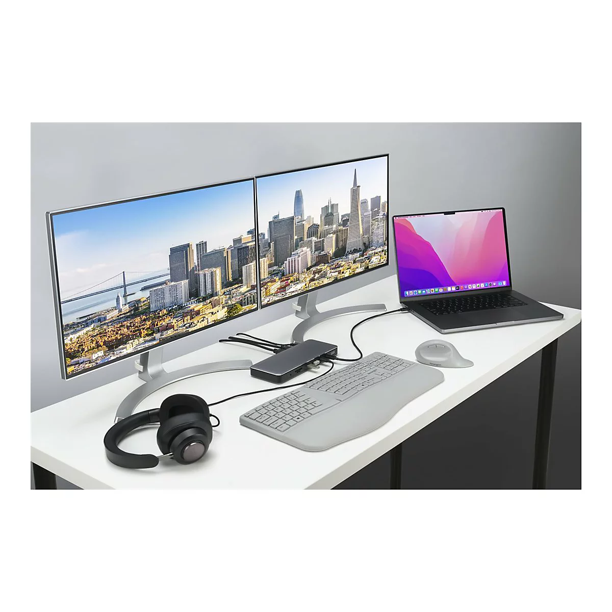 Schreibtisch mit zwei Monitoren, Laptop, Tastatur, Maus und KopfhÃ¶rern. Auf den Monitoren ist eine Stadtansicht zu sehen.