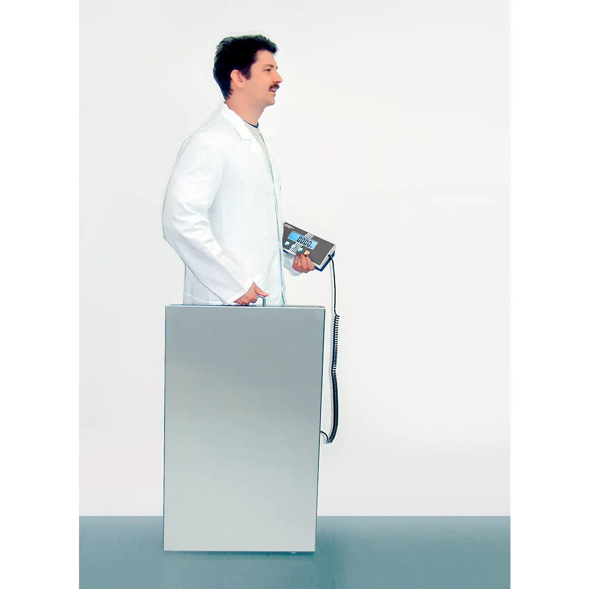 Un homme en blouse blanche se tient derrière une armoire métallique, tenant un appareil avec un câble.