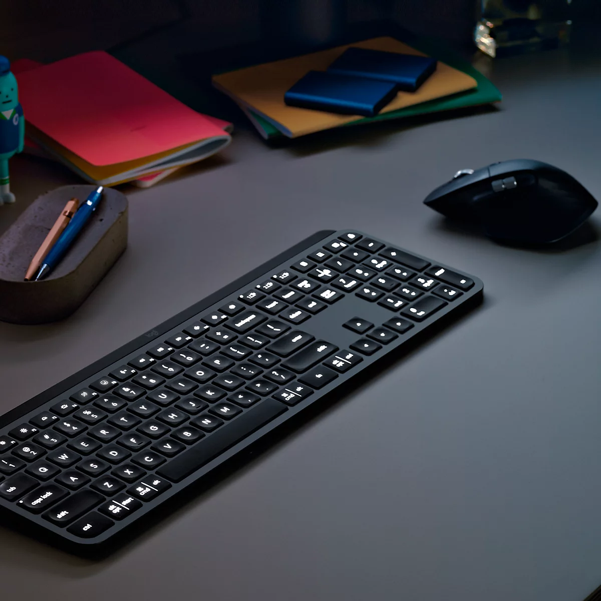 Black illuminated keyboard on a grey desk. A mouse on the right, with notes and pens next to it.
