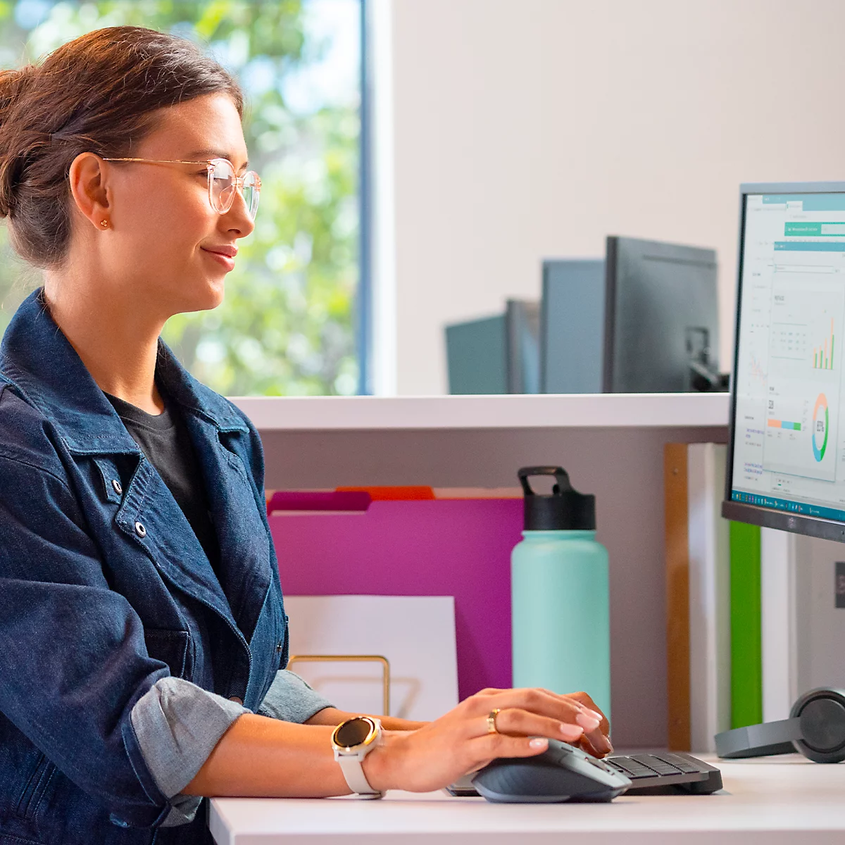 A woman sits at a desk, working on a computer and smiling. She wears glasses and a denim jacket. On the table: water bottle, folders.