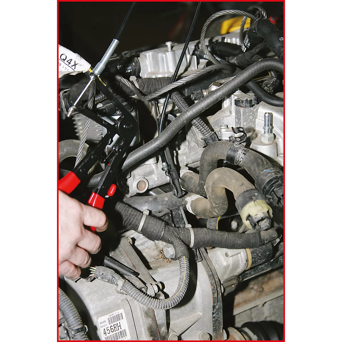 Hände halten eine Zange an einem Motor. Rote Griffe, schwarzer Zangenkopf.