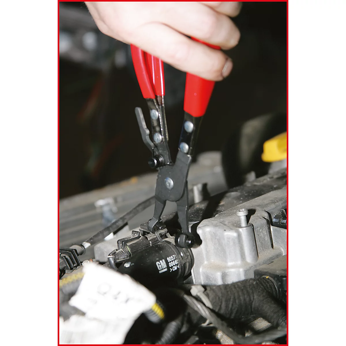 Eine Hand bedient eine Zange an einem Motor. Die Zange hat rote Griffe, der Motor ist teilweise zu sehen.