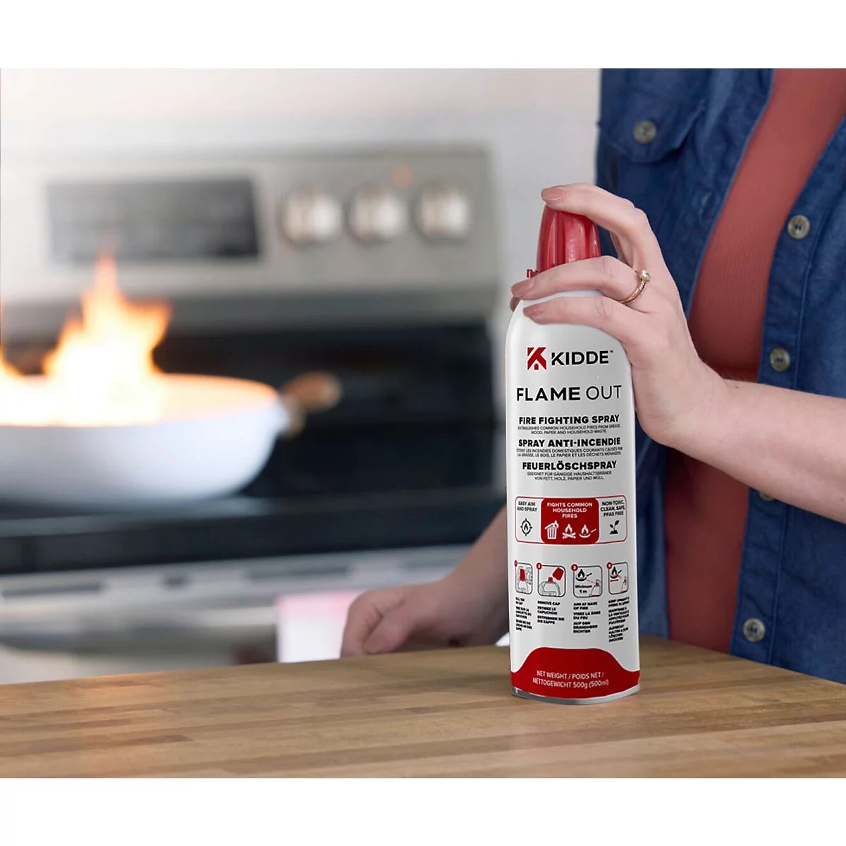 Person hält eine Kidde Flame Out Feuerlöschspray-Dose in der Hand, während im Hintergrund eine Pfanne auf dem Herd brennt.