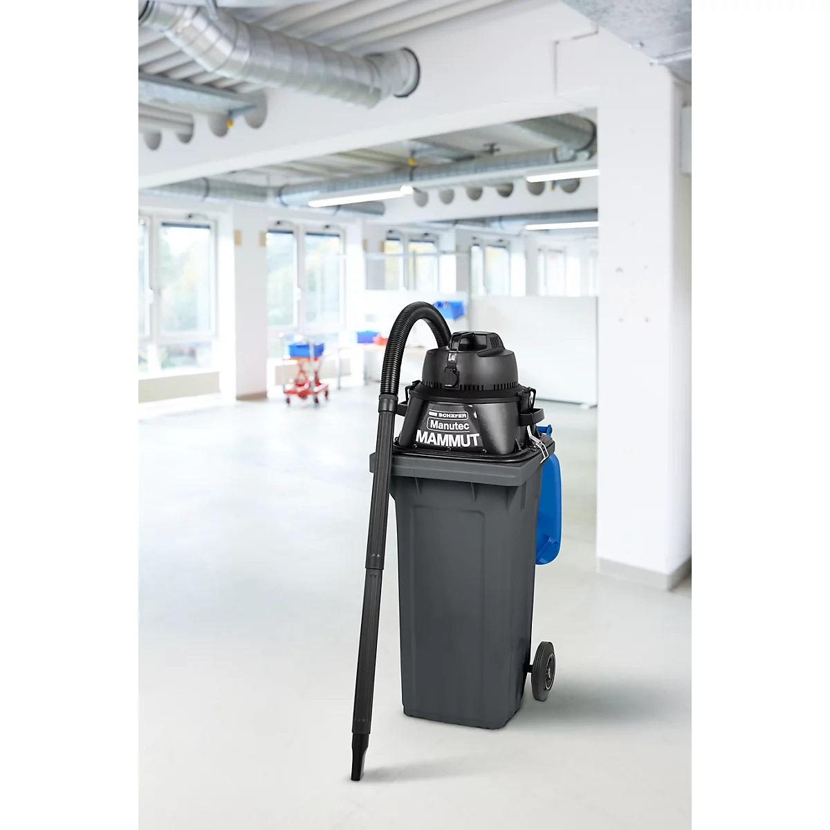 Aspirateur industriel noir sur conteneur à déchets gris, devant un bureau lumineux. L'aspirateur porte l'inscription Mammut.