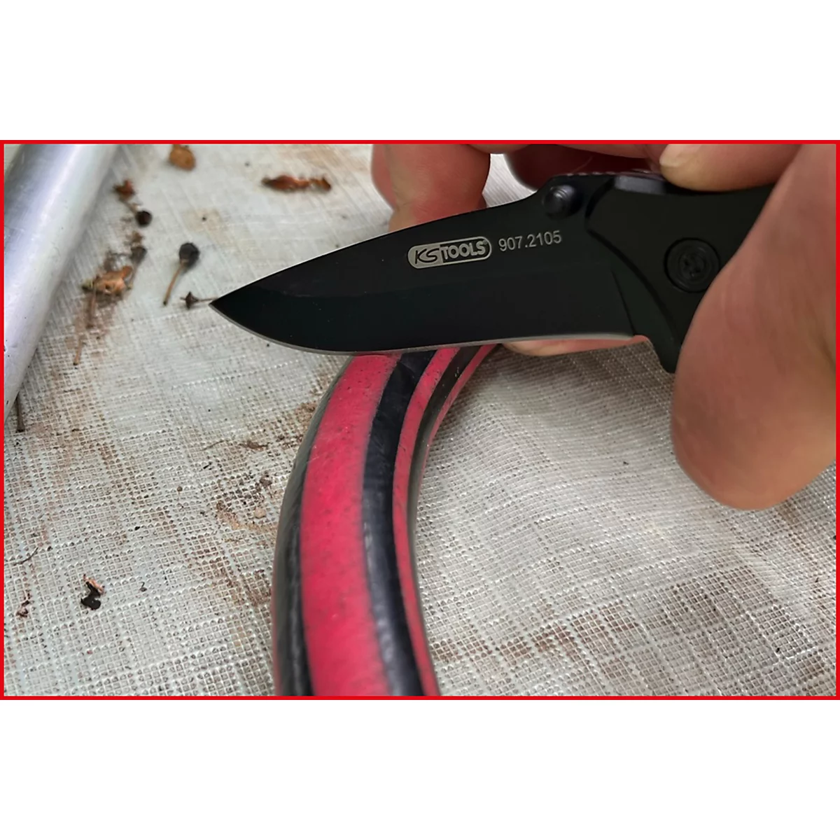 Schwarzes Klappmesser schneidet einen roten Schlauch mit schwarzen Streifen. Auf der Klinge steht 'KS TOOLS 907.2105'.