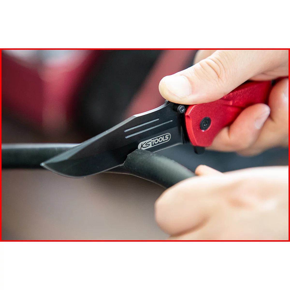Nahaufnahme eines roten Klappmessers, das ein schwarzes Kabel durchschneidet. Auf der Klinge steht 'KSTOOLS'.