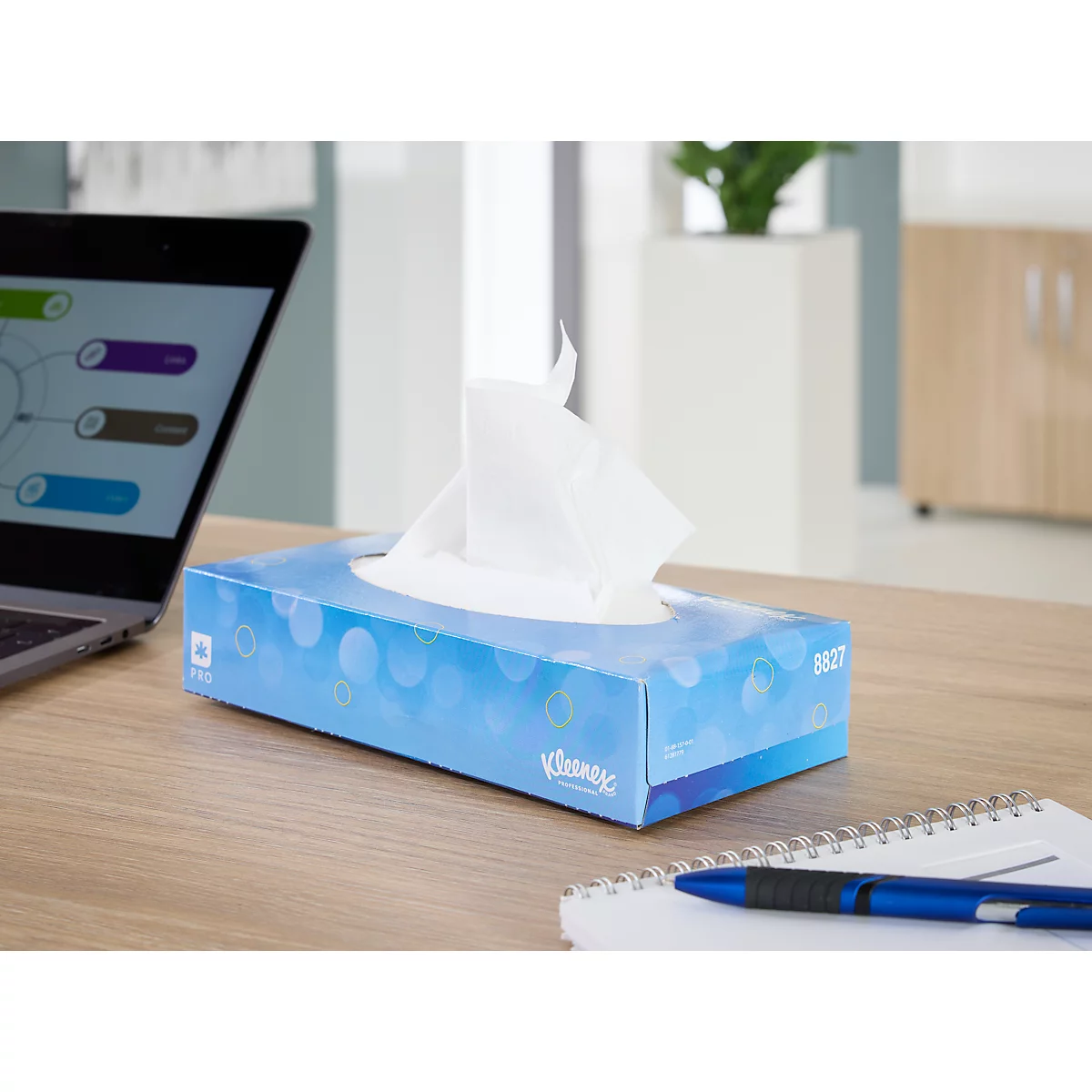 Een blauwe Kleenex-doos op een bureau. Daarnaast een laptop, notitieblok en pen.
