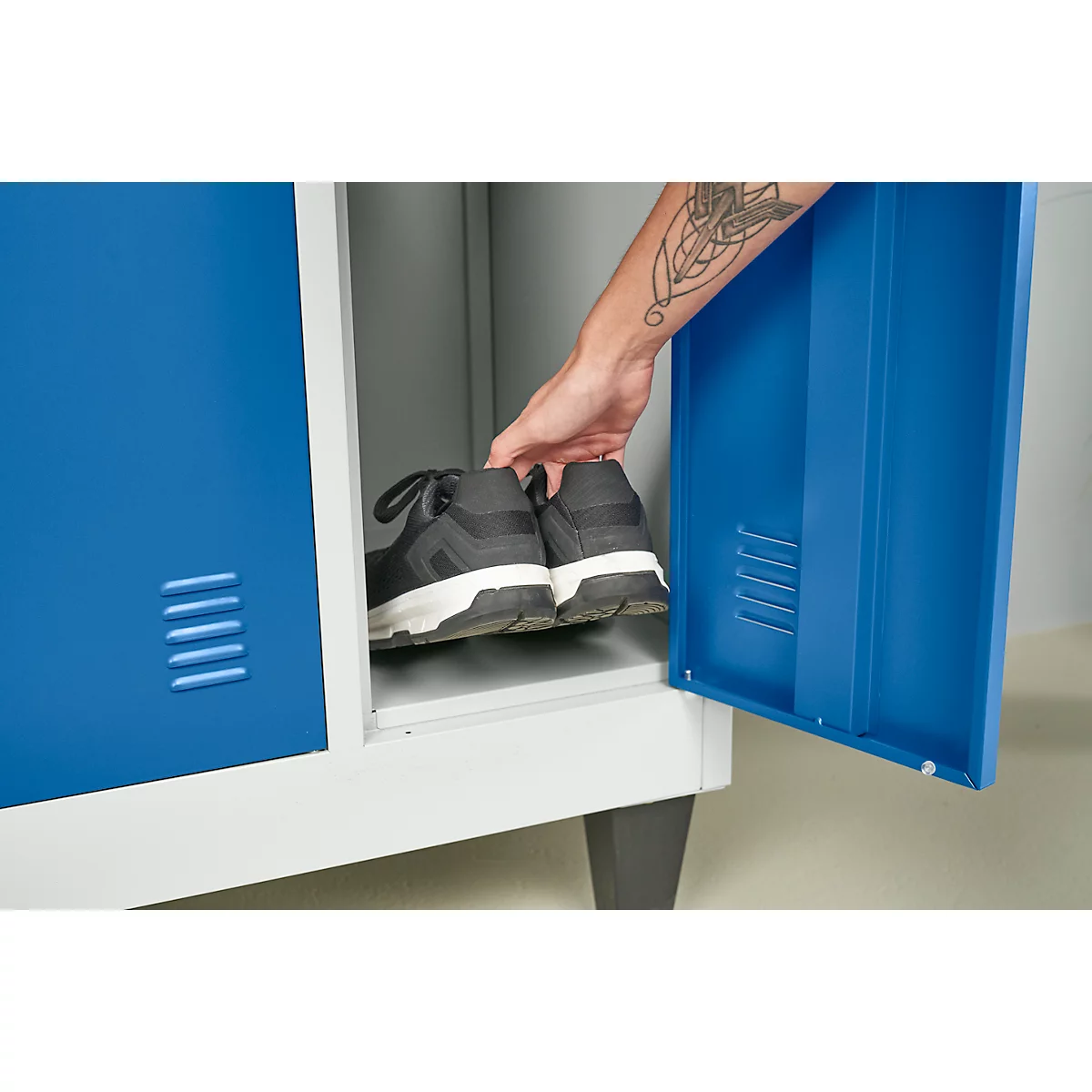 Hand legt Schuhe in ein blaues Schließfach. Dahinter graue Wände und ein grauer Boden.