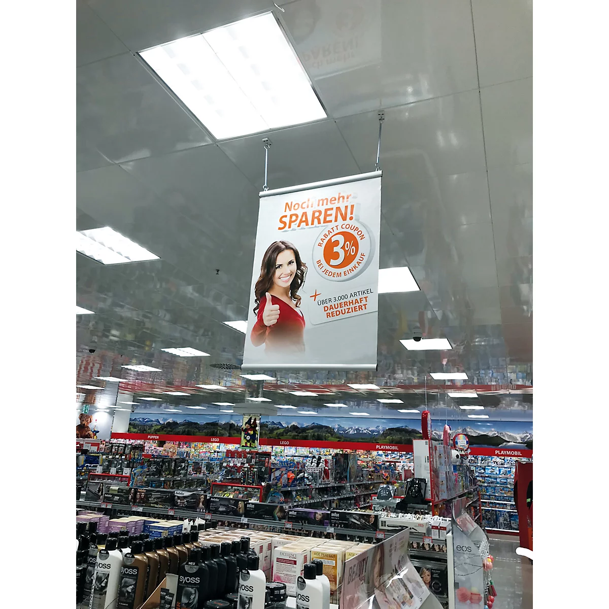 Ein Banner im Geschäft wirbt für 3% Rabatt. Eine Frau mit Daumen hoch lächelt. Der Text lautet: Noch mehr sparen!