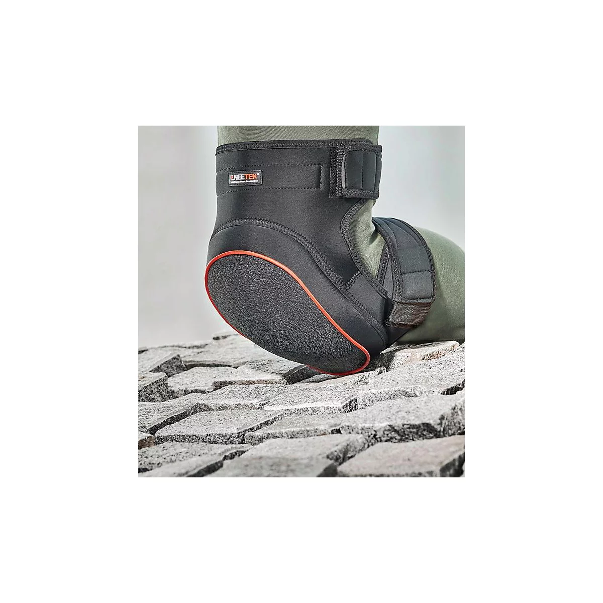 Genouillère noire avec bordure rouge, sur sol en pierre, jambe dans un pantalon vert olive.