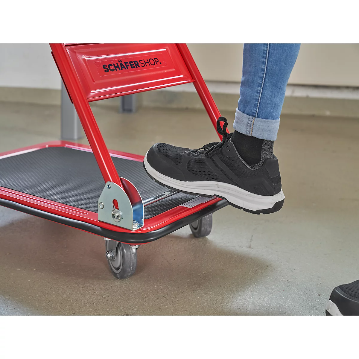 Nahaufnahme eines roten Transportwagens mit einem Fuß, der die Plattform betätigt. Der Schriftzug „SCHÄFERSHOP“ ist zu sehen.